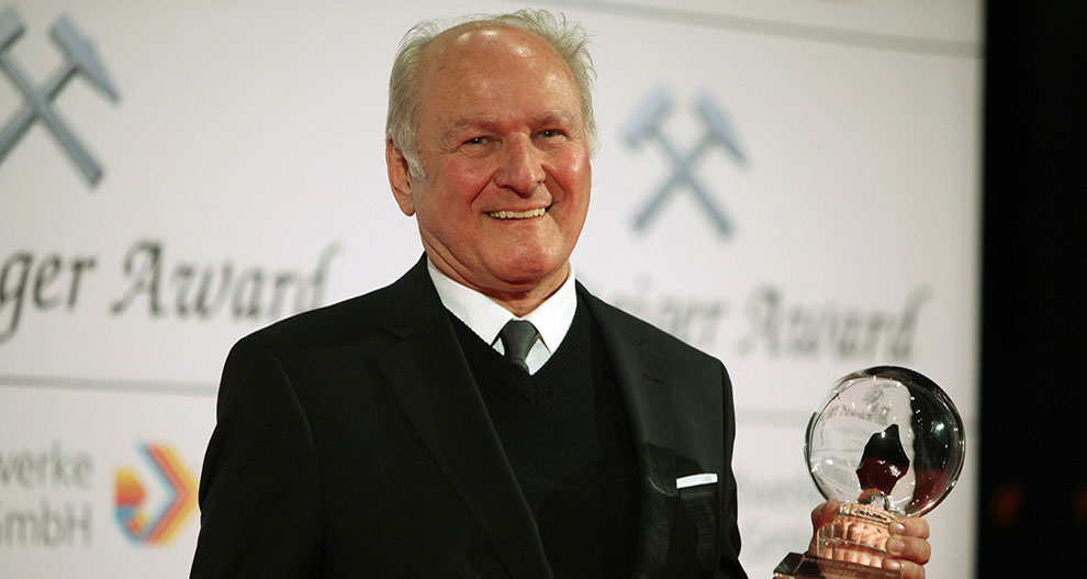 An elderly man in a black suit stands on a stage, holding a transparent spherical award, with the words "Schweizer Award" and a logo visible in the background.
