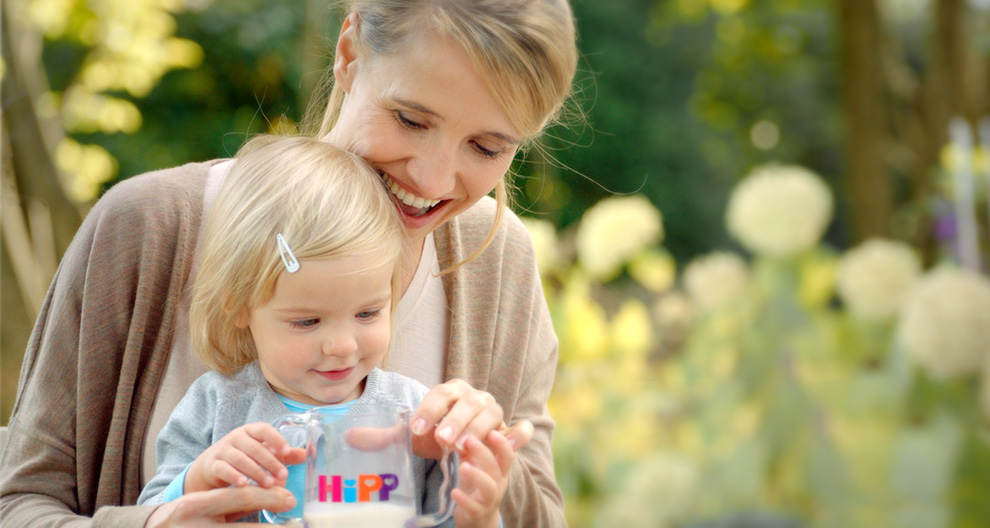 A woman and a young child are outdoors, looking at a clear container with the word "HiPP" written on it.