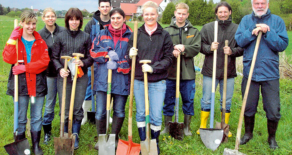 A group of people stands in a grassy field holding shovels, ready for a gardening or construction task.